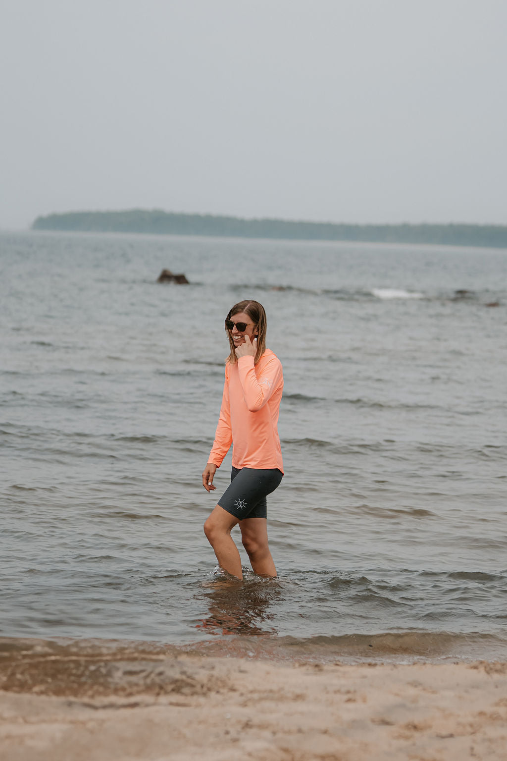 woman walking in shallow water on a beach in the coral colour UPF 50+ hooded long sleeve shirt and grey UPF 50+ swim shorts