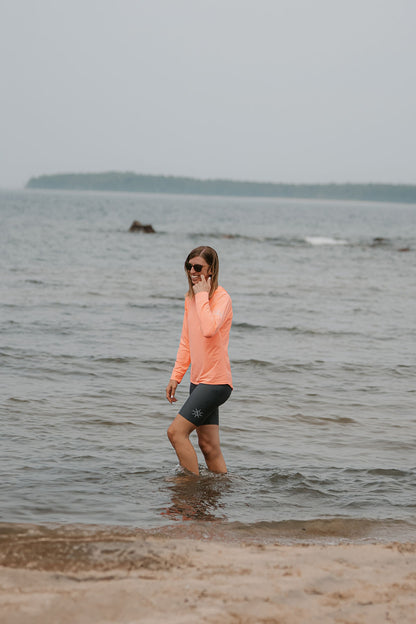 woman walking in shallow water on a beach in the coral colour UPF 50+ hooded long sleeve shirt and grey UPF 50+ swim shorts