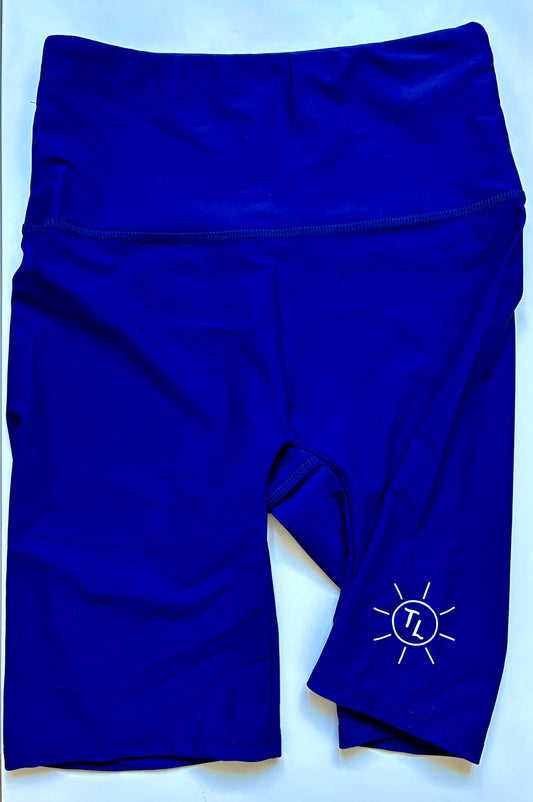 A pair of navy blue women's UPF 50+ swim shorts with a high waist and above knee-length fit, featuring a small white logo on the lower left leg.